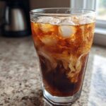 Close-up of a tall glass filled with an Iced Vanilla Chai Latte, showing ice cubes and cream swirling into the dark tea.