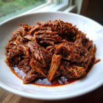 Close-up of tender, shredded Instant Pot Beef Barbacoa coated in rich, dark red sauce, served in a white bowl.