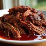 Close-up of shredded, saucy Instant Pot Beef Barbacoa piled high on a white plate, ready for tacos.