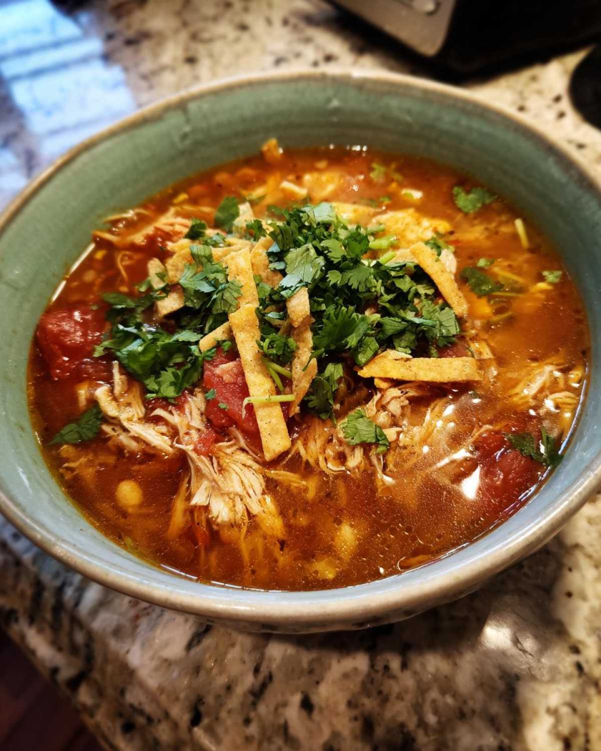 Close-up of a bowl of rich, red broth Instant Pot Chicken Tortilla Soup topped with shredded chicken, tomatoes, tortilla strips, and fresh cilantro.