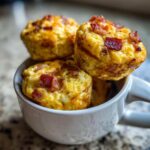 Three golden Keto Bacon and Egg Muffins stacked inside a white mug, topped with crispy bacon bits.