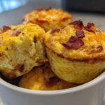 A stack of golden Keto Bacon and Egg Muffins topped with melted cheese and bacon bits, served in a white mug.