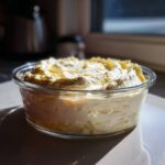 A glass bowl filled with fluffy, pale yellow Keto Lemon Cheesecake Mousse, highlighted by sunlight.