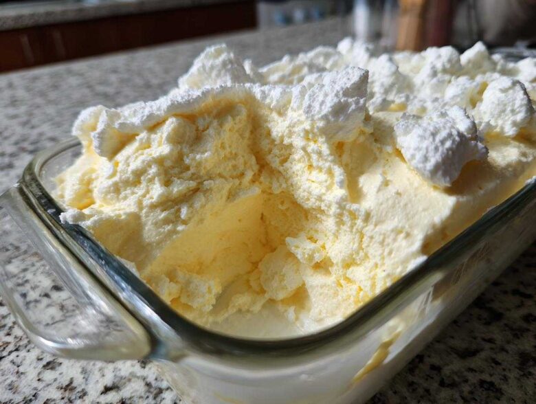 Close-up of creamy, pale yellow Keto Lemon Cheesecake Mousse topped with dollops of white whipped topping in a glass dish.