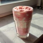 A tall glass filled with a swirled pink and white Keto Strawberry Cheesecake Smoothie, sitting on a marble counter near a window.