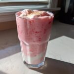 A tall glass filled with a pink Keto Strawberry Cheesecake Smoothie, topped with whipped cream and cinnamon.