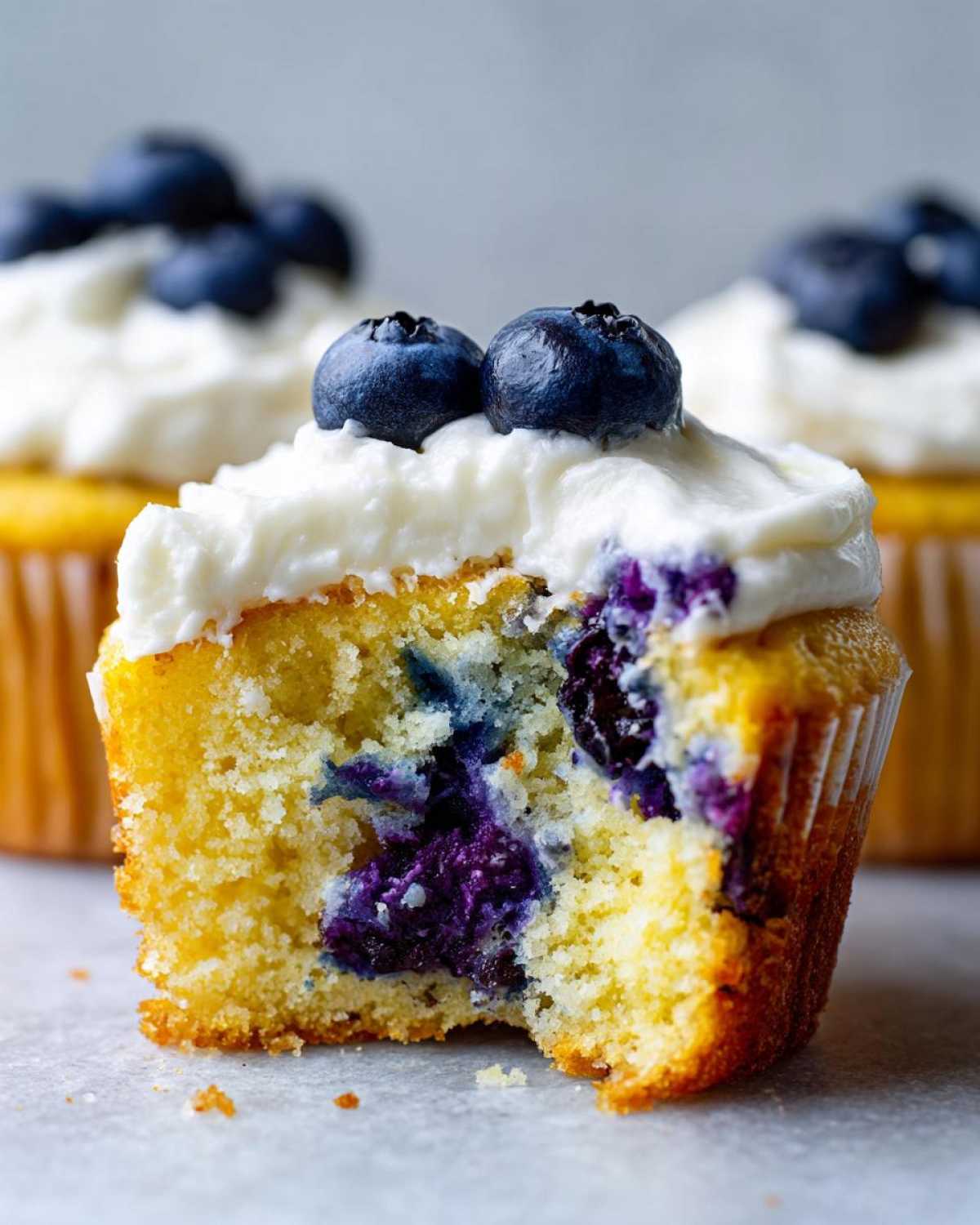 Close-up of a Lemon Blueberry Cupcake with cream cheese frosting, showing a bite taken out revealing moist cake and burst blueberries.