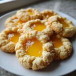 A close-up view of several crumbly Lemon Curd Thumbprint Cookies filled with bright yellow lemon curd on a white plate.
