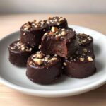 A stack of rich, dark chocolate covered banana bites topped with chopped nuts, one showing a creamy interior.