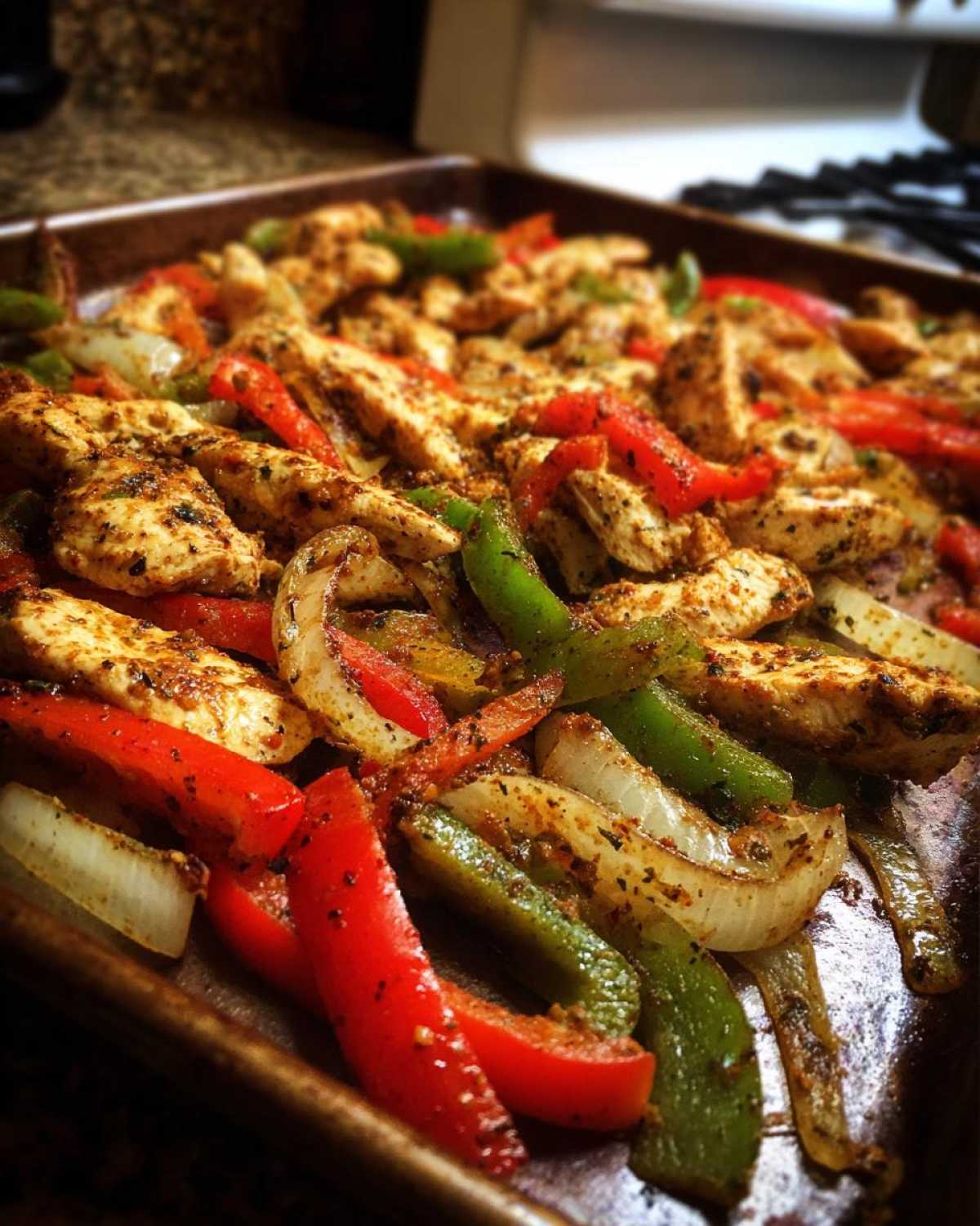Close-up of seasoned chicken strips mixed with sliced red peppers, green peppers, and onions cooked on a sheet pan for One-Pan Chicken Fajitas.