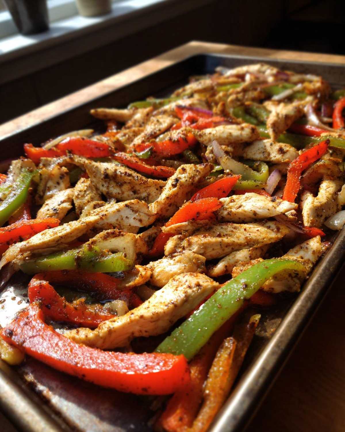 Close-up of seasoned chicken strips mixed with sliced red and green peppers and onions on a dark sheet pan for One-Pan Chicken Fajitas.