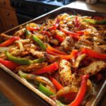 Close-up of seasoned chicken strips, red bell peppers, green peppers, and onions cooked on a sheet pan for One-Pan Chicken Fajitas with Peppers and Onions.
