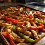 Close-up of seasoned chicken strips, red bell peppers, green bell peppers, and onions cooked together on a sheet pan for One-Pan Chicken Fajitas with Peppers and Onions.