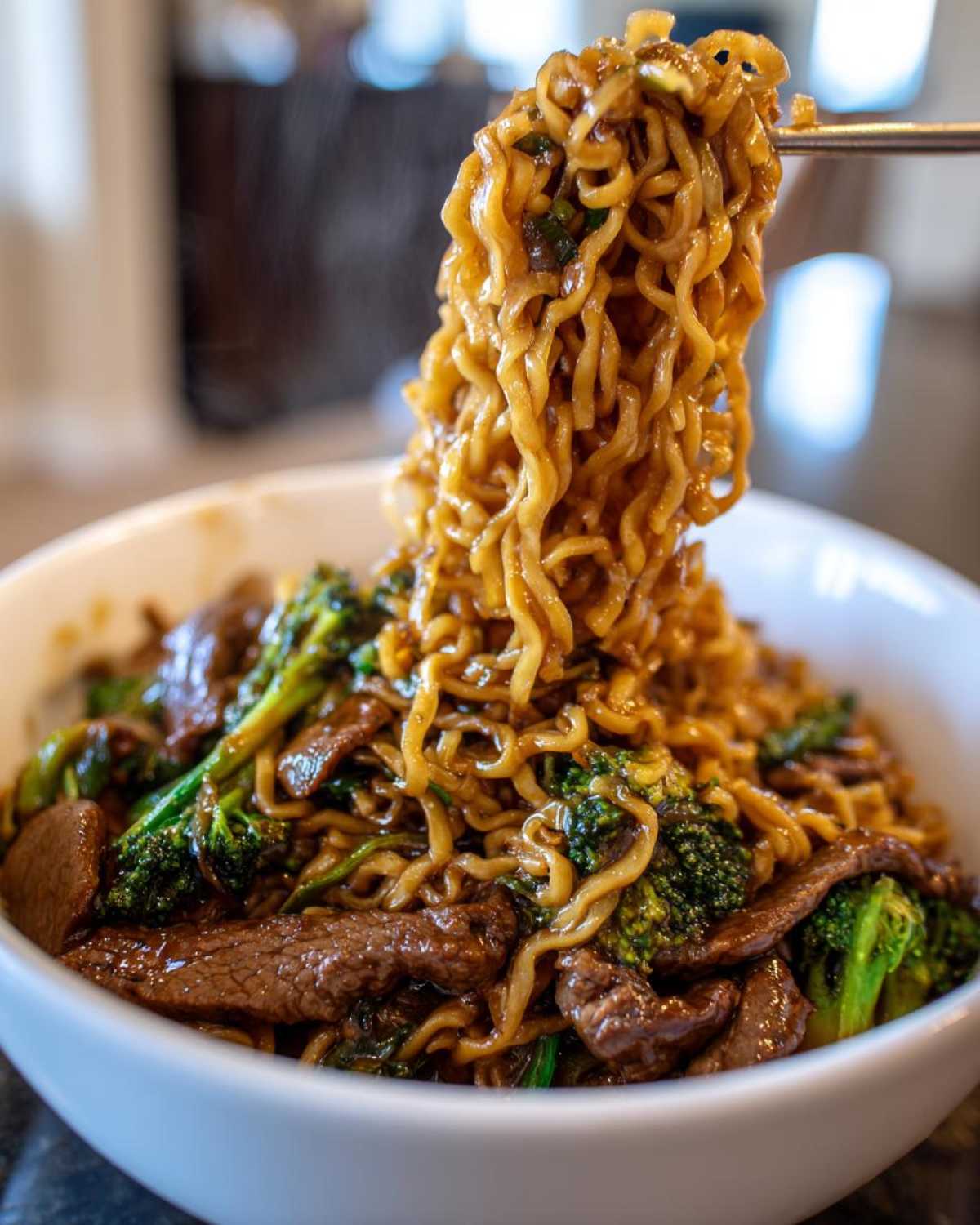 A close-up shot of steaming One-Pot Beef and Broccoli Ramen Noodles being lifted from a white bowl with chopsticks.