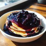 A stack of fluffy pancakes generously covered with thick Homemade Blueberry Syrup in a white bowl.