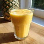 A vibrant yellow Pineapple Coconut Smoothie served in a tall glass on a wooden counter, with a whole pineapple in the background.