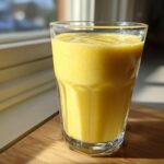 A tall glass filled with a vibrant yellow Pineapple Coconut Smoothie sitting on a wooden surface near a window.