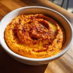 A close-up of vibrant orange Roasted Red Pepper Hummus swirled with olive oil and sprinkled with paprika in a white bowl.