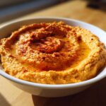 A close-up of creamy Roasted Red Pepper Hummus swirled in a white bowl, drizzled with olive oil and sprinkled with paprika.
