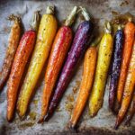 Close-up of glossy Sheet Pan Roasted Rainbow Carrots with Honey, showing orange, yellow, and purple varieties on parchment paper.