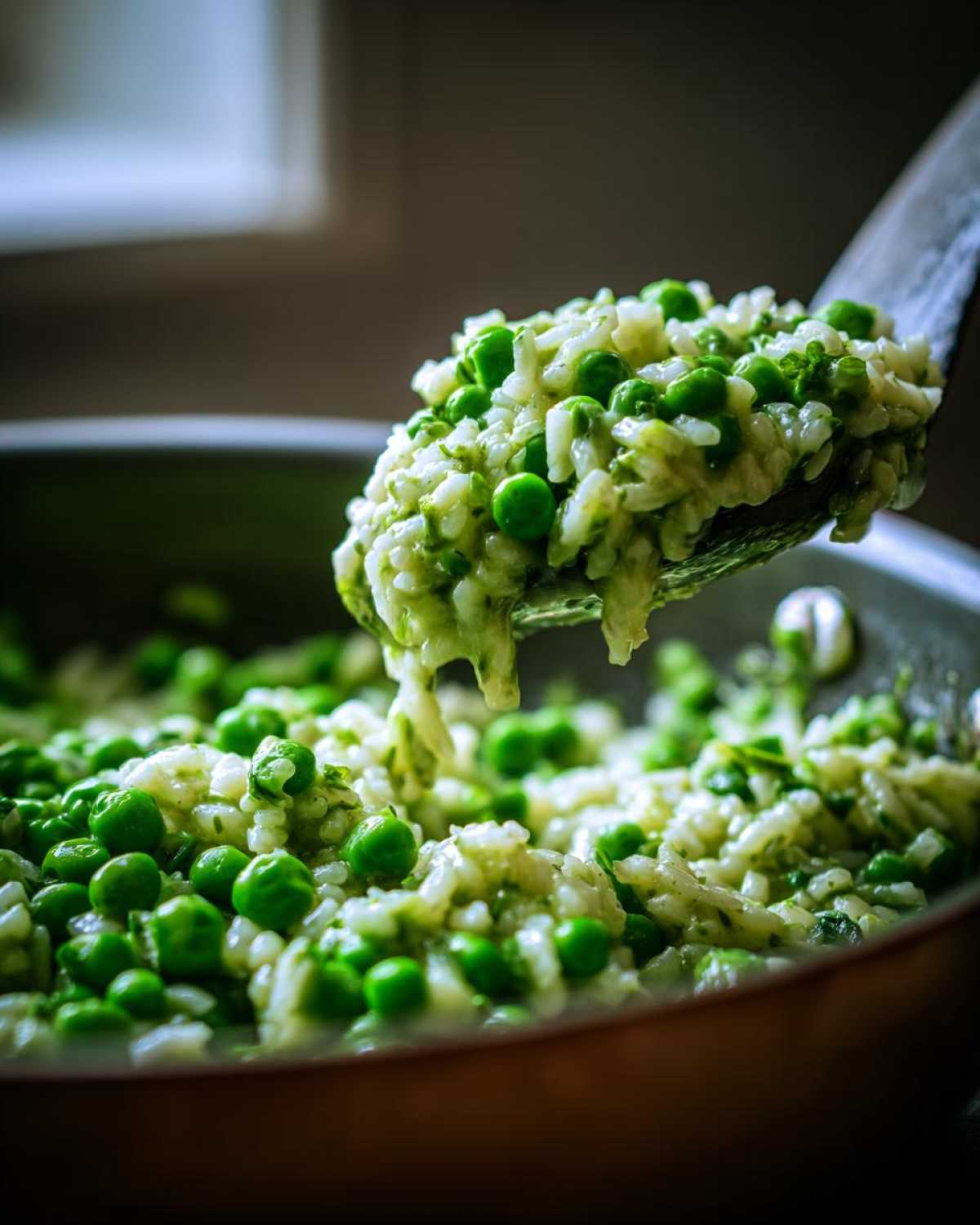 A wooden spoon lifts a creamy portion of Spring Pea and Mint Risotto, showing bright green peas mixed with rice, out of a cooking pan.