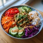 A vibrant Spring Roll Bowl with Peanut Sauce featuring rice noodles, shredded carrots, cucumber, red cabbage, and protein.