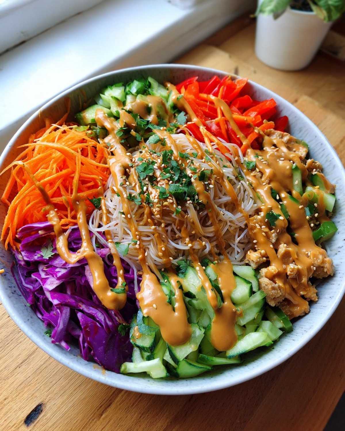 A vibrant Spring Roll Bowl with Peanut Sauce featuring rice noodles, shredded carrots, red cabbage, cucumbers, and bell peppers.