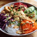 A close-up of a colorful Spring Roll Bowl with Peanut Sauce, featuring vermicelli noodles, shredded carrots, cabbage, and cucumber.