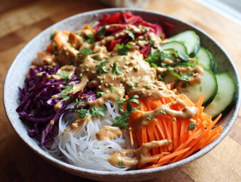 A close-up of a colorful Spring Roll Bowl with Peanut Sauce, featuring vermicelli noodles, shredded carrots, cabbage, and cucumber.