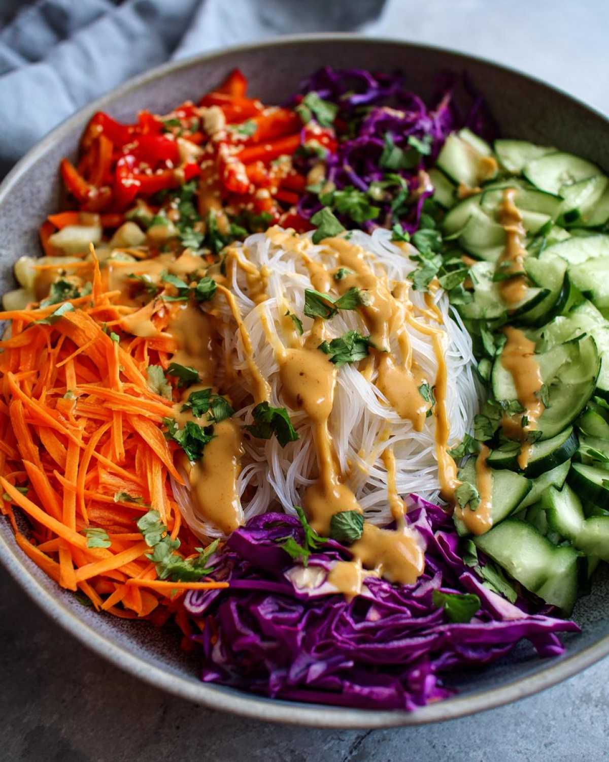 A colorful Spring Roll Bowl with rice noodles, shredded carrots, cucumbers, red cabbage, and drizzled peanut sauce.
