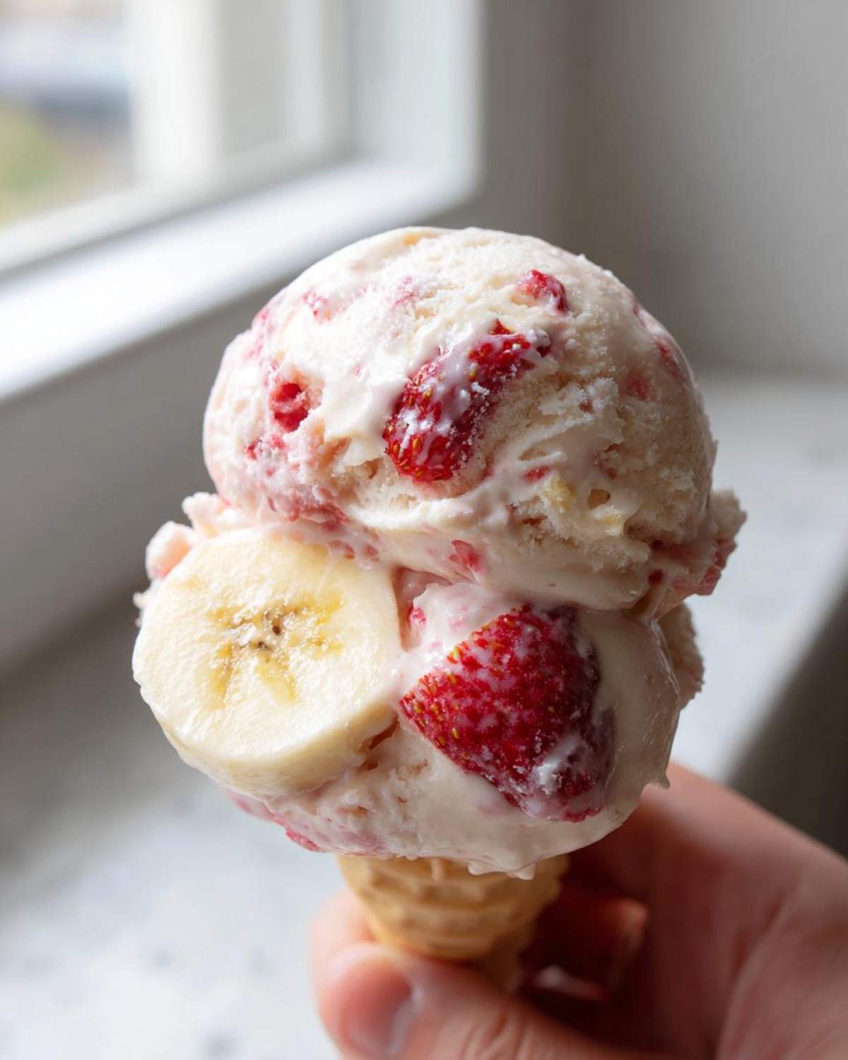 Close-up of two scoops of Strawberry Banana Ice Cream in a waffle cone, featuring visible chunks of strawberries and banana slices.