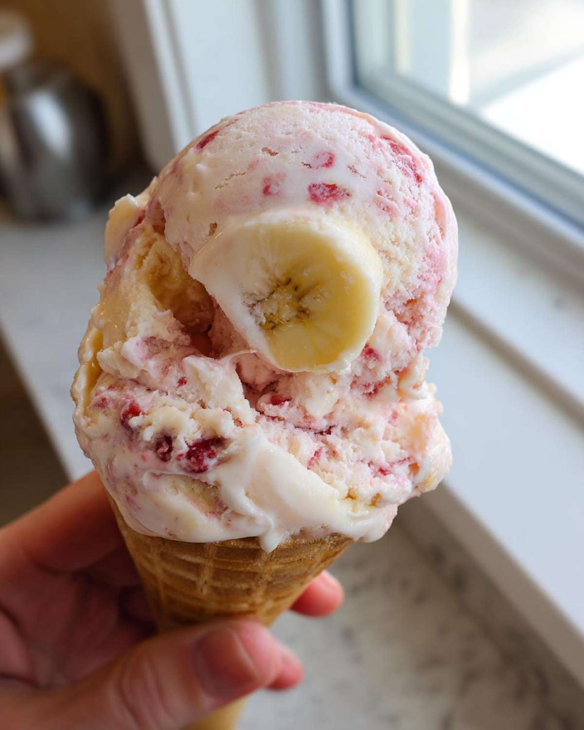 A close-up of a scoop of pink and white Strawberry Banana Ice Cream topped with a banana slice, held in a waffle cone.