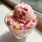 Three scoops of pink and yellow Strawberry Banana Ice Cream swirled with fruit pieces in a clear glass dish.