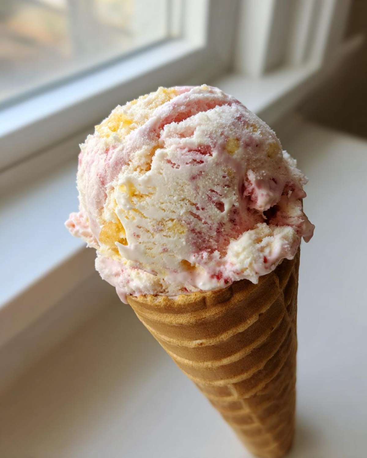 A perfect scoop of swirled Strawberry Banana Ice Cream served in a crisp waffle cone, set against a bright window.