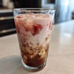 A tall glass filled with layered Strawberry Cream Cold Brew, showing coffee, cream, and strawberry pieces over ice.