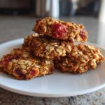 A stack of four golden brown Strawberry Oatmeal Breakfast Cookies featuring visible oats and pieces of red strawberry on a white plate.