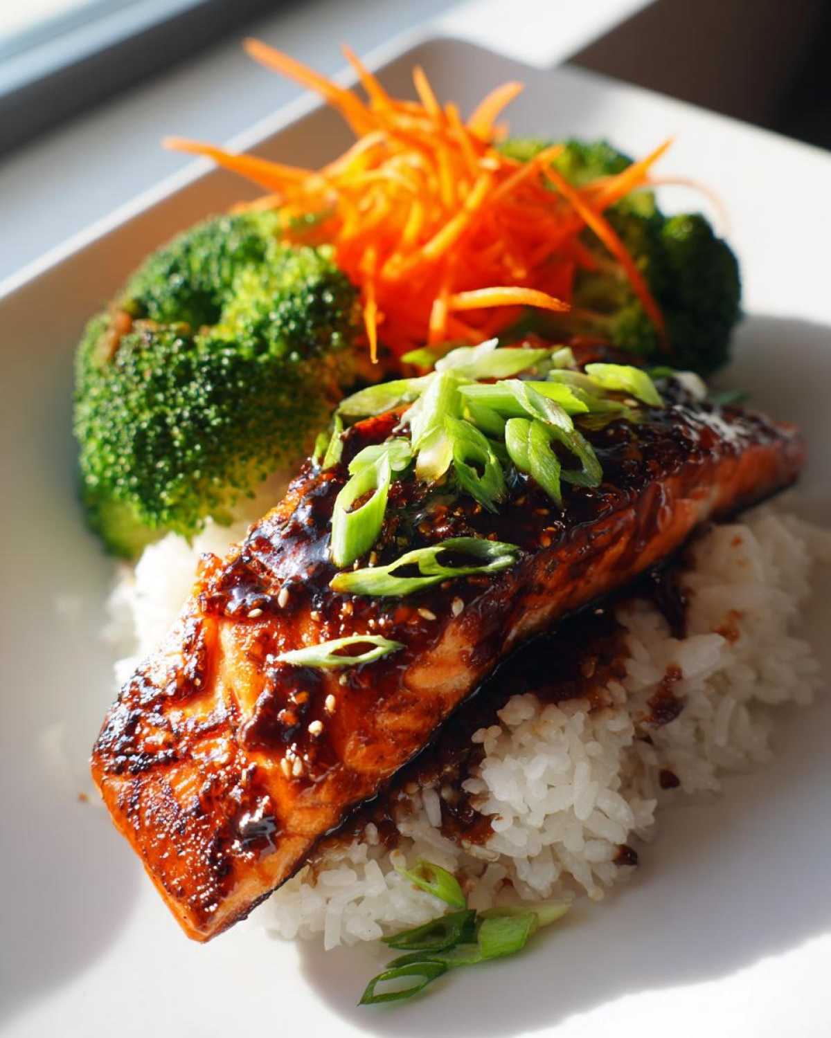 A piece of glazed Teriyaki Salmon served over white rice, garnished with green onions, broccoli, and shredded carrots.
