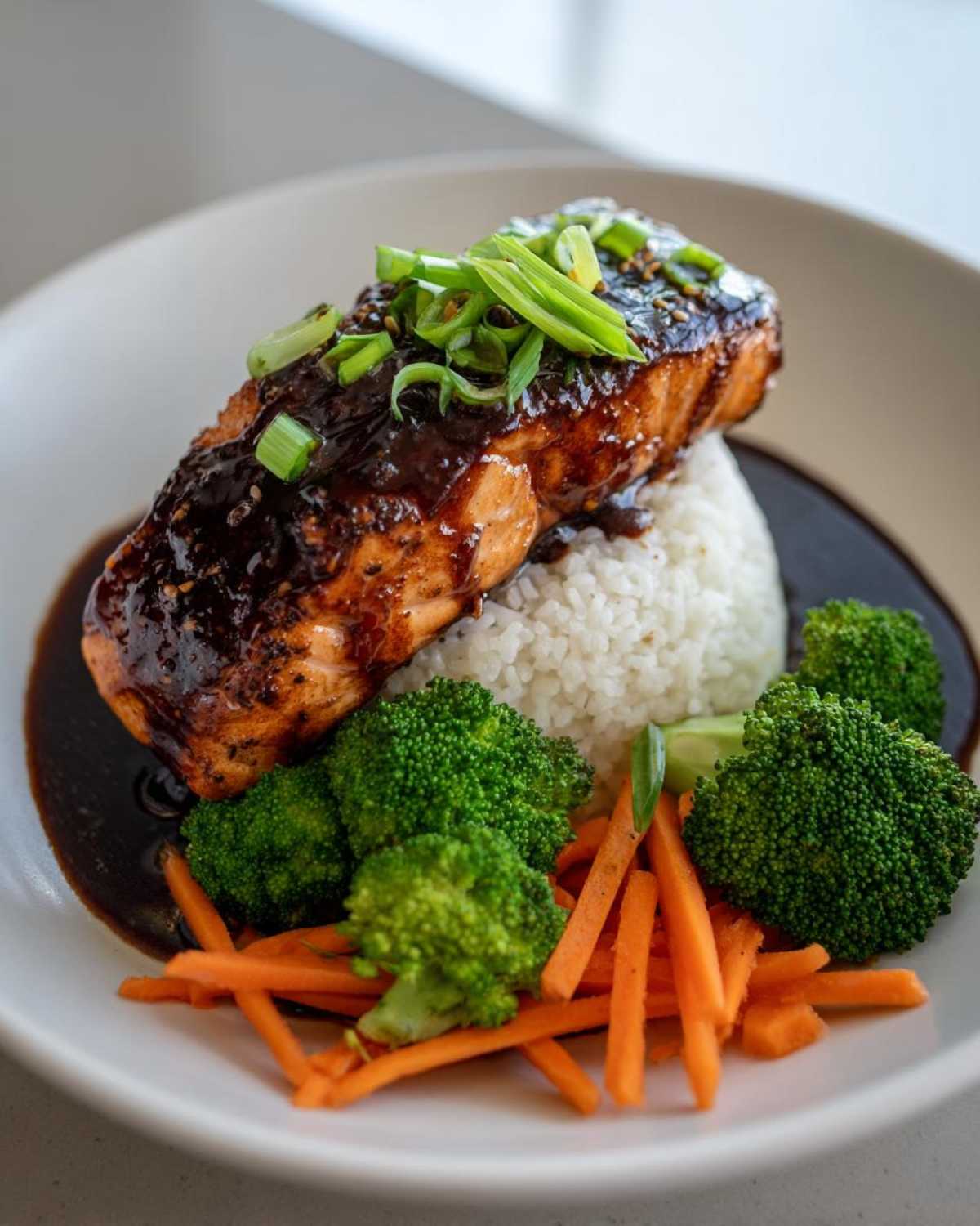 A beautifully plated Teriyaki Salmon Rice Bowls featuring a thick piece of glazed salmon over white rice, served with steamed broccoli and julienned carrots.