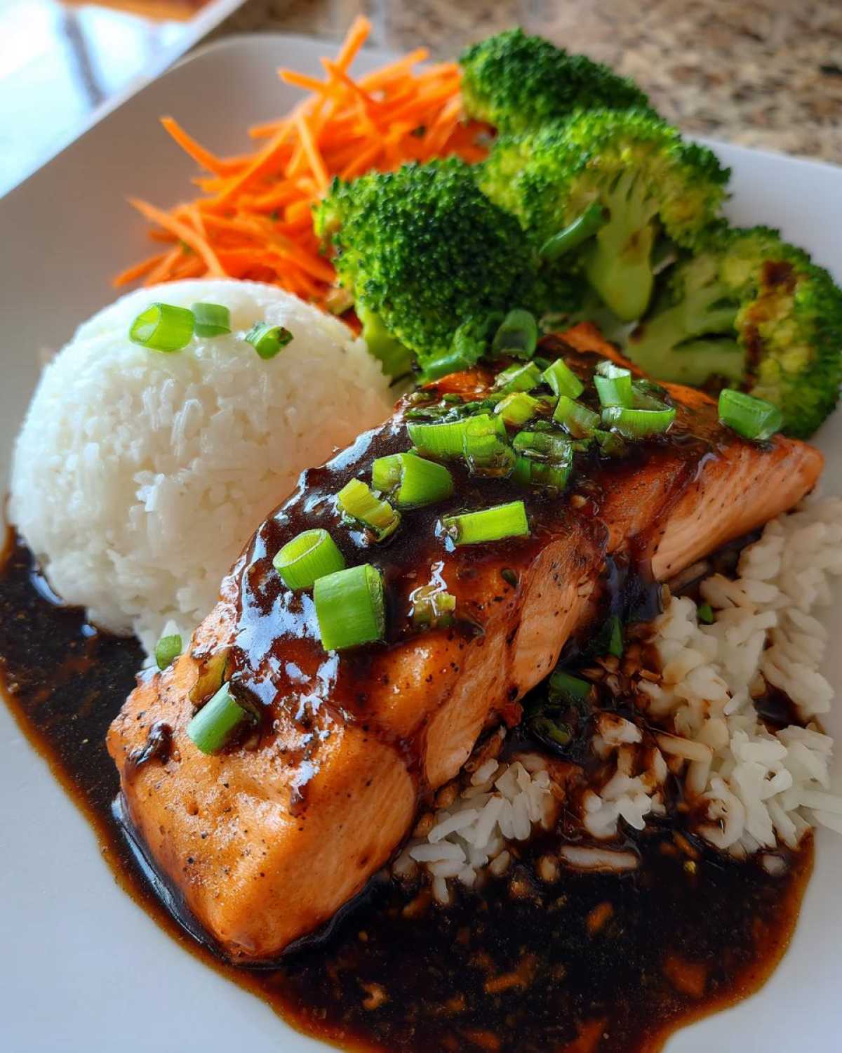A close-up of a serving of Teriyaki Salmon Rice Bowls featuring glazed salmon over white rice, broccoli, and shredded carrots.