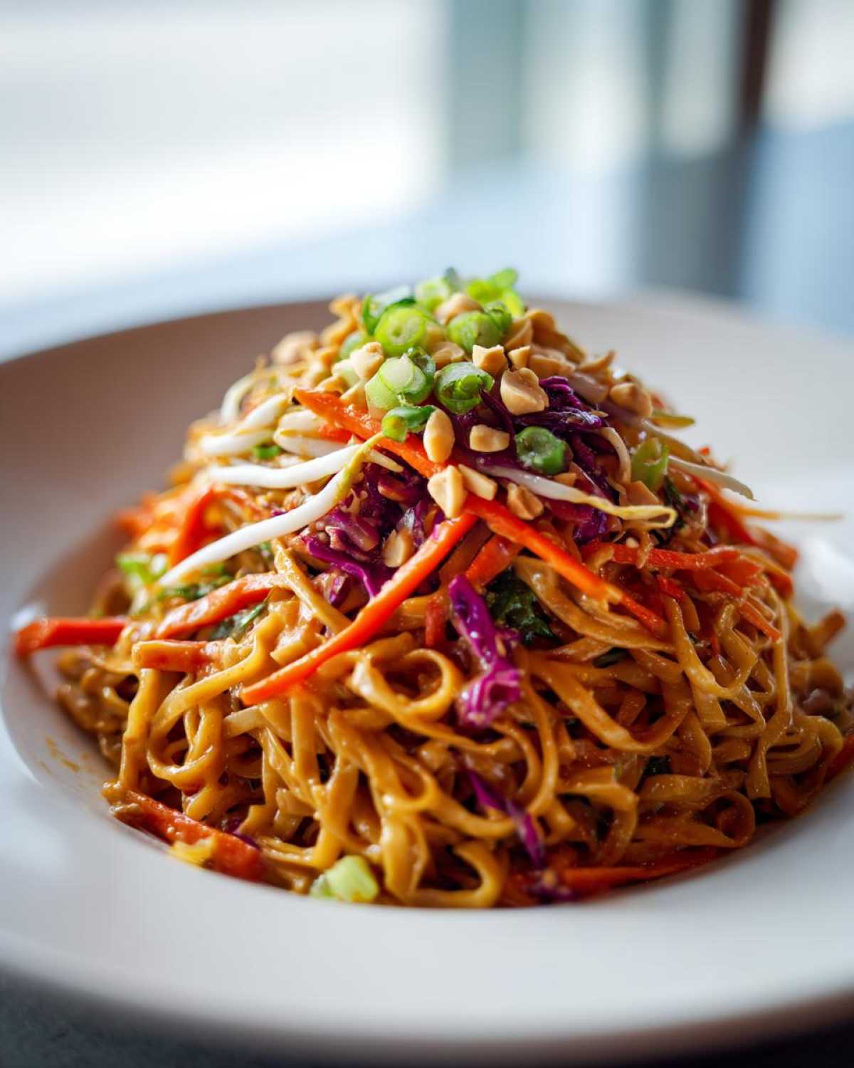A vibrant bowl of Thai Peanut Noodles with crunchy shredded carrots, red cabbage, bean sprouts, and chopped peanuts.