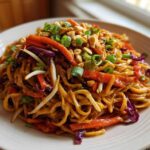 A close-up of a plate piled high with saucy Thai Peanut Noodles with Crunchy Veggies, topped with peanuts and scallions.