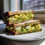 Two halves of an Ultimate Avocado Egg Salad Sandwich stacked on a white plate, showing creamy green filling and egg chunks.