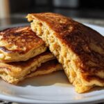 A stack of golden-brown, fluffy Vegan Banana Oat Pancakes drizzled with syrup on a white plate.