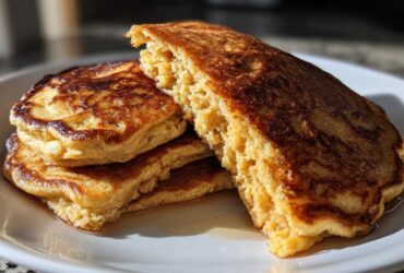 A stack of golden-brown, fluffy Vegan Banana Oat Pancakes drizzled with syrup on a white plate.