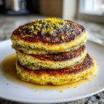 A stack of three golden Vegan Lemon Poppy Seed Pancakes dotted with poppy seeds and topped with bright lemon zest.