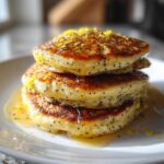 A close-up of three fluffy Vegan Lemon Poppy Seed Pancakes stacked and drizzled with syrup and lemon zest.