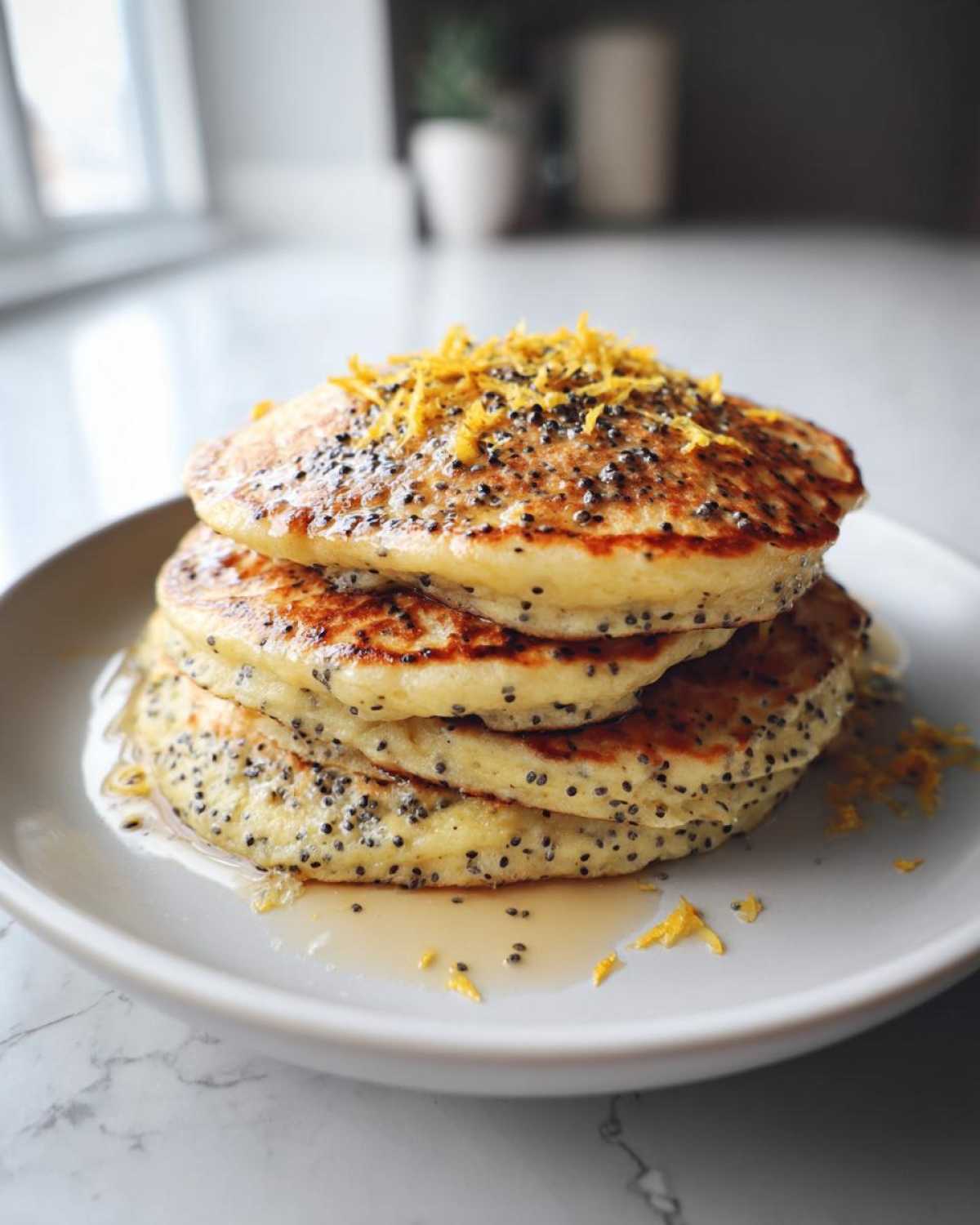 A tall stack of golden Vegan Lemon Poppy Seed Pancakes dotted with poppy seeds, drizzled with syrup and topped with bright lemon zest.