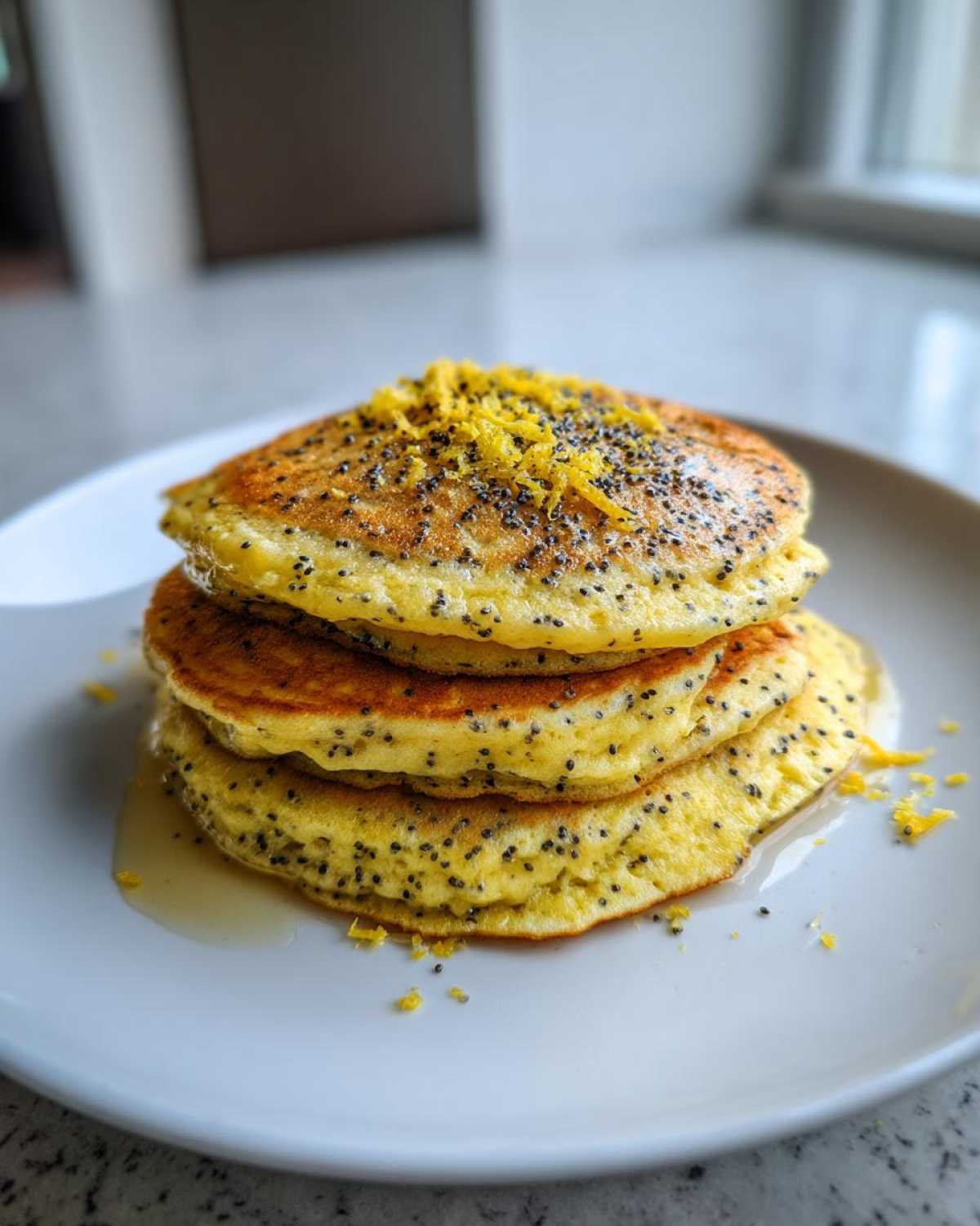 A stack of three fluffy Vegan Lemon Poppy Seed Pancakes topped with lemon zest and drizzled with syrup.