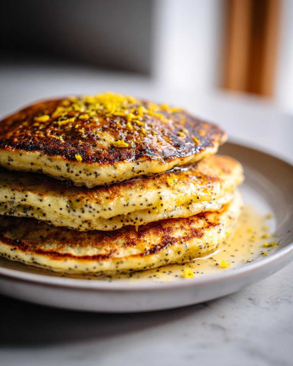 A stack of three golden Vegan Lemon Poppy Seed Pancakes drizzled with syrup and topped with bright yellow lemon zest.