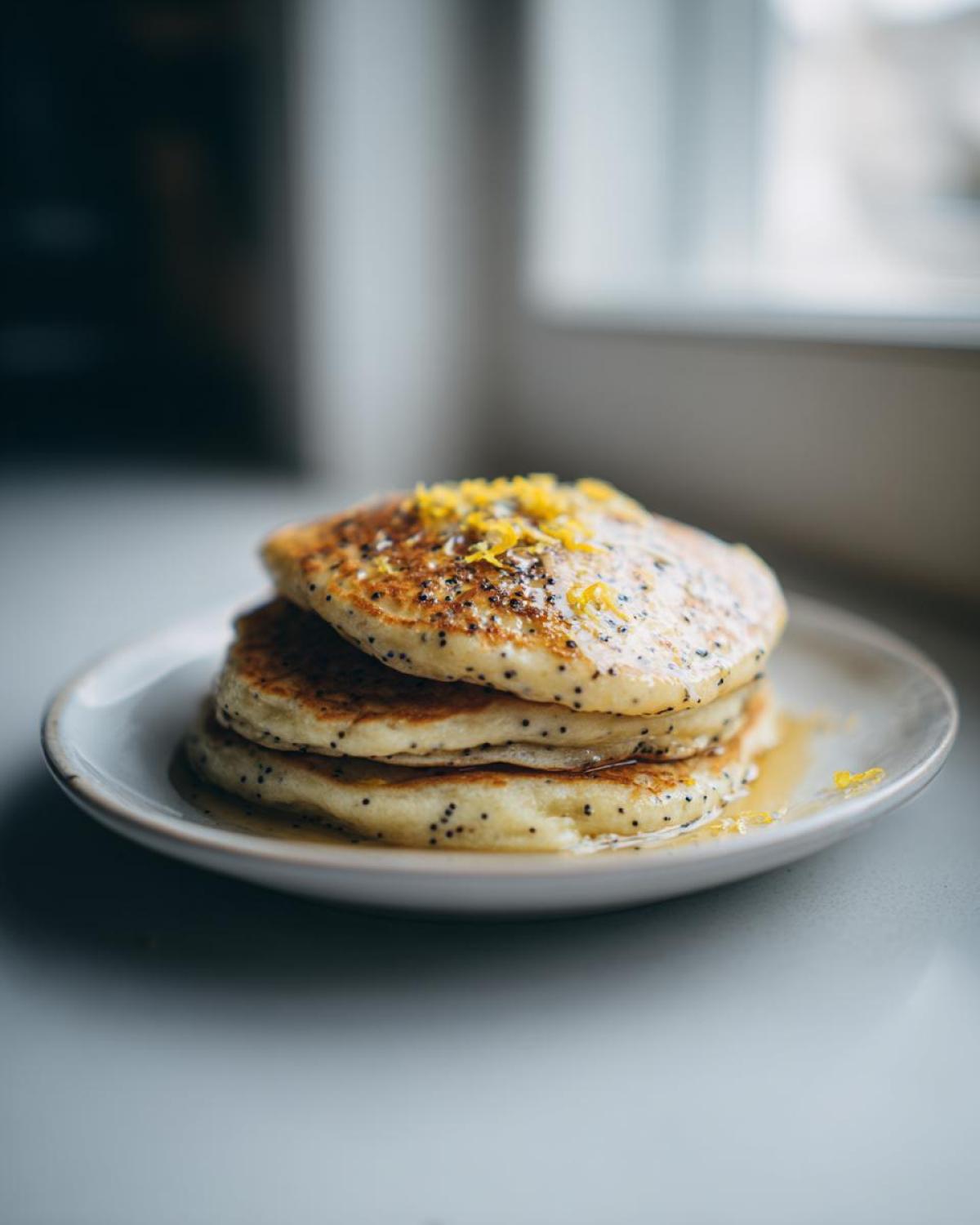 A stack of three fluffy Vegan Lemon Poppy Seed Pancakes topped with lemon zest and syrup.